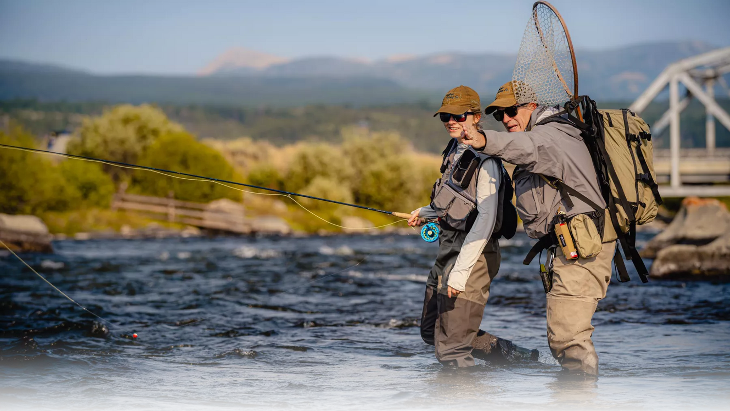Two people fly fishing in a river with mountains in the background